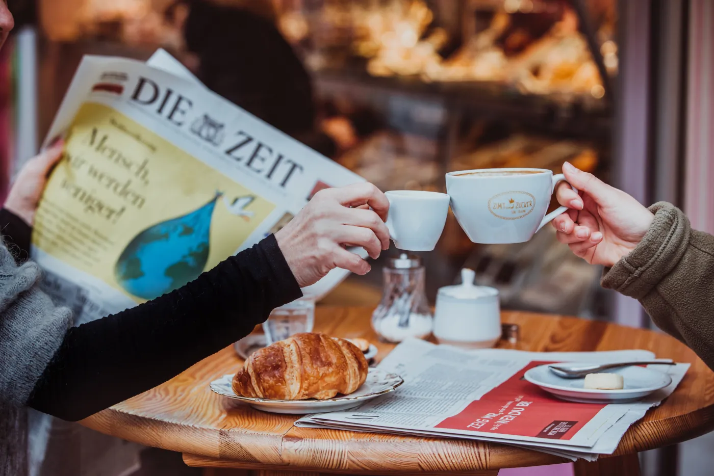 Zwei Personen stoßen mit Zimt-und-Zucker-Kaffeetassen an einem Cafétisch mit Zeitung und Croissant an