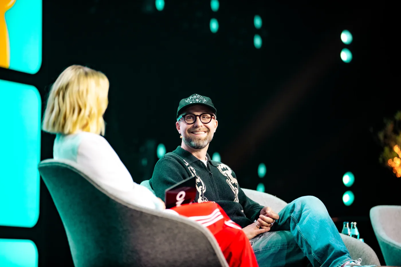 Mann mit Cap und Brille lächelt im Bühnengespräch neben einer Interviewerin, im Hintergrund bunte Bühnenlichter