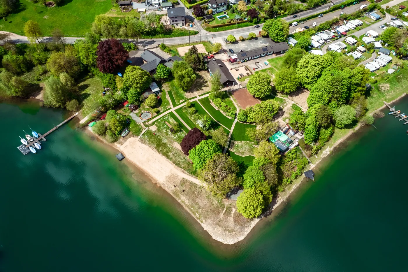 Luftaufnahme einer grünen Halbinsel an einem See mit Gartenwegen, Bootssteg und angrenzendem Campingplatz
