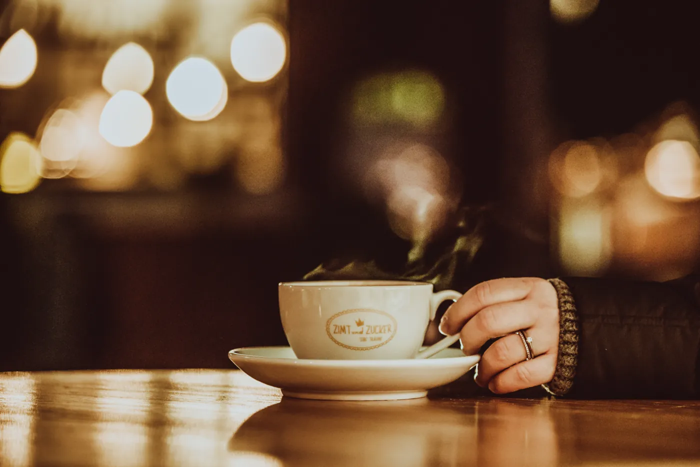 Hand haelt eine Kaffeetasse mit Zimt-und-Zucker-Logo in warmer Caféatmosphäre