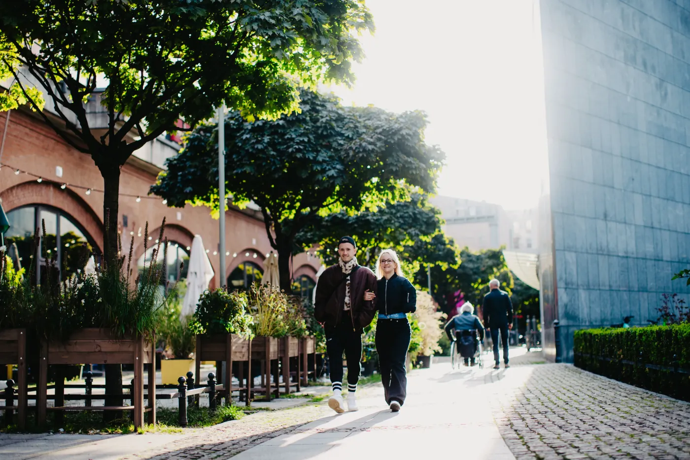 Paar geht Arm in Arm eine sonnige Promenade entlang, im Hintergrund Passanten und eine Rollstuhlfahrerin
