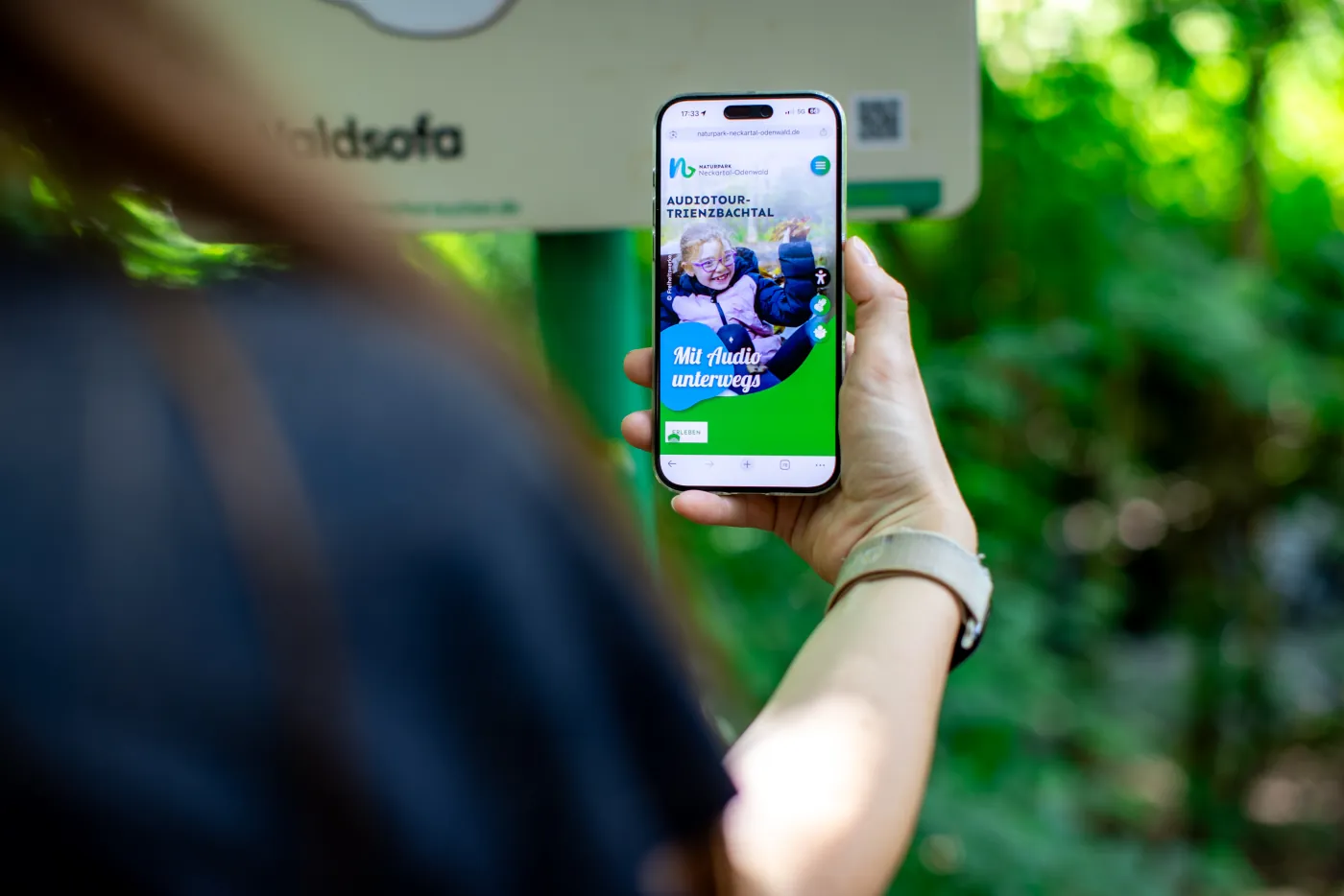 Person hält ein Smartphone mit einer Audiotour-Webseite vor einem Wegweiser im Wald